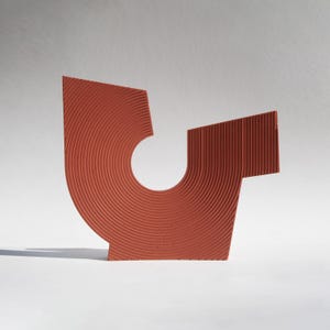 以下が含まれることがあります： テラコッタ色の抽象彫刻。円形の切り抜きとテクスチャ加工された表面が特徴です。 彫刻は、曲線と直線で構成された幾何学的なデザインで、白い背景に影を落としています。