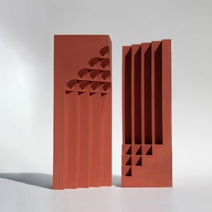Può includere: Due sculture rettangolari color terracotta con disegni geometrici. Una presenta intagli ad arco e linee verticali, mentre l'altra ha scanalature verticali e intagli triangolari. Le sculture sono su sfondo bianco.