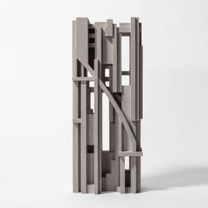Op de afbeelding: Een hoge, rechthoekige sculptuur van lichtgrijs, gespikkeld materiaal met een complexe opstelling van verticale en horizontale staven, kruisende balken en open rechthoekige ruimtes, wat een gelaagde, architecturale en geometrische structuur vormt.