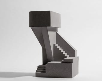 Puede incluir: Una escultura arquitectónica gris que presenta una serie de bloques geométricos y escaleras interconectados. La estructura incluye una plataforma elevada prominente apoyada por un pilar central, con múltiples juegos de escaleras a distintos niveles.