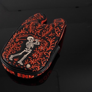 May include: A black golf putter headcover featuring an intricate orange floral vine pattern and a central graphic of a skeleton swinging a golf club. The side of the headcover displays the red text "PUTTER END" in a bold, capitalised font.