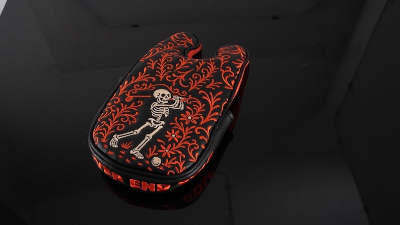 May include: A black golf putter headcover featuring an intricate orange floral vine pattern and a central graphic of a skeleton swinging a golf club. The side of the headcover displays the red text "PUTTER END" in a bold, capitalised font.