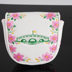 May include: A white mallet putter headcover featuring an embroidered green arched bridge with a yellow flag at the center. The design is framed by pink cherry blossom flowers with yellow centers and green stems, stitched onto the white surface.