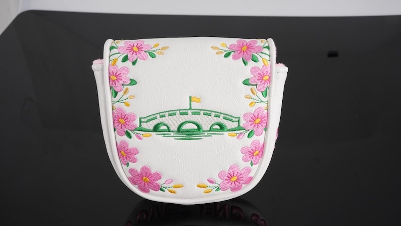 May include: A white mallet putter headcover featuring an embroidered green arched bridge with a yellow flag at the center. The design is framed by pink cherry blossom flowers with yellow centers and green stems, stitched onto the white surface.