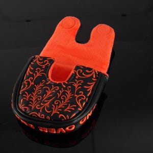 May include: A black golf putter headcover featuring an orange filigree pattern on the top surface. The interior is lined with bright orange fabric, and the exterior edge displays orange text that reads OVER AND OUT in a bold, capitalized font.