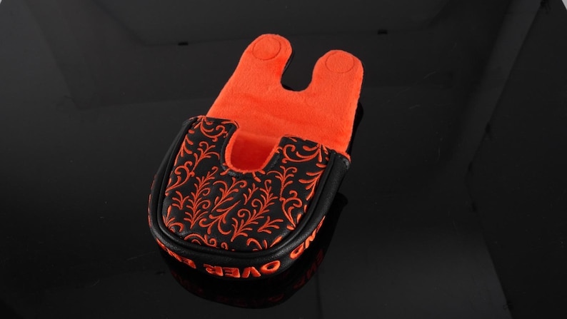 May include: A black golf putter headcover featuring an orange filigree pattern on the top surface. The interior is lined with bright orange fabric, and the exterior edge displays orange text that reads OVER AND OUT in a bold, capitalized font.