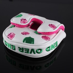 Puede incluir: Una funda blanca para putter de golf con flores rosas y hojas verdes bordadas. El lateral de la funda muestra el texto END OVER END en letras verdes llamativas. El forro interior y el borde son de un color rosa intenso y vibrante.