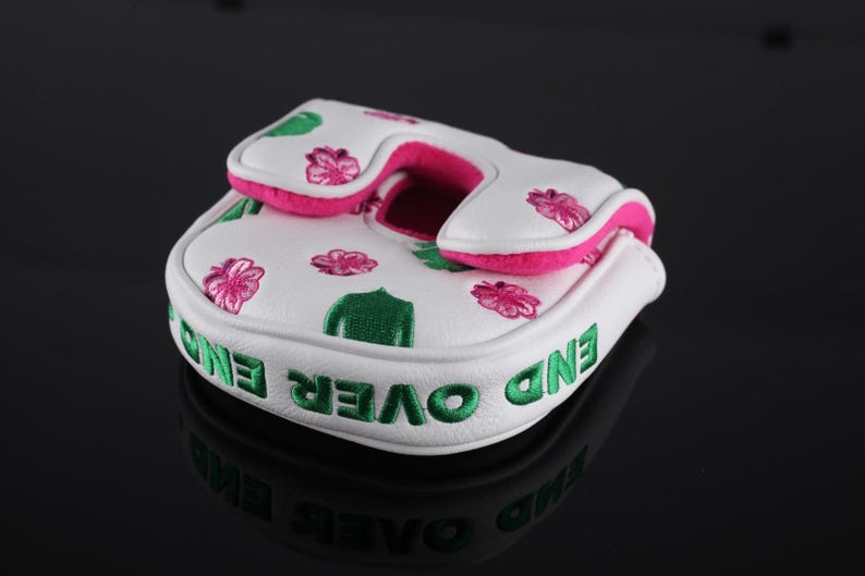 Puede incluir: Una funda blanca para putter de golf con flores rosas y hojas verdes bordadas. El lateral de la funda muestra el texto END OVER END en letras verdes llamativas. El forro interior y el borde son de un color rosa intenso y vibrante.