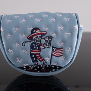 May include: A light blue golf putter cover with white stars and a skeleton golfer design. The skeleton wears a patriotic outfit and holds a golf club. An American flag is also embroidered on the cover.