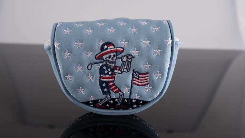 May include: A light blue golf putter cover with white stars and a skeleton golfer design. The skeleton wears a patriotic outfit and holds a golf club. An American flag is also embroidered on the cover.