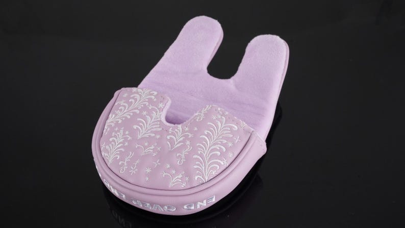 May include: A lavender golf putter head cover with a U-shaped top and a decorative floral pattern in white. The cover has the words "END OVER" printed on the bottom edge. The background is black.