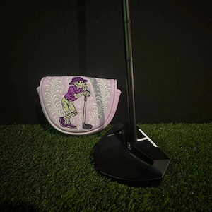 May include: Black golf putter with a white 'T' marking and a matching putter cover. The cover is lavender with a skeleton graphic in purple, green, and white. The putter and cover are on green turf.