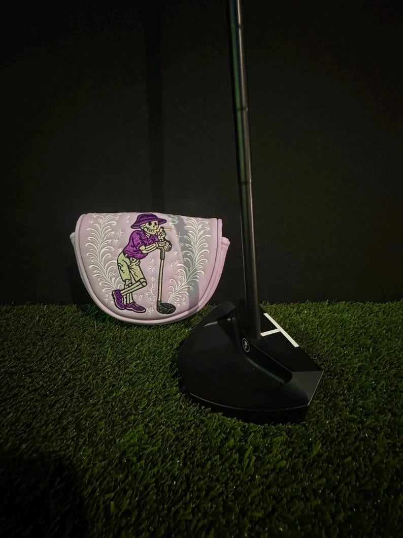 May include: Black golf putter with a white 'T' marking and a matching putter cover. The cover is lavender with a skeleton graphic in purple, green, and white. The putter and cover are on green turf.