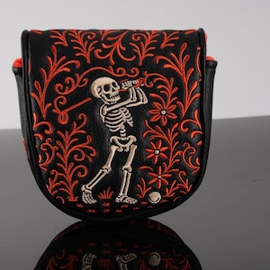 May include: A black mallet-style golf putter headcover featuring an embroidered white skeleton swinging a golf club. The design is surrounded by intricate orange floral and vine patterns that extend across the front of the leather-like cover.