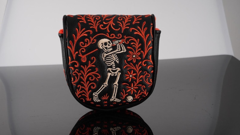 May include: A black mallet-style golf putter headcover featuring an embroidered white skeleton swinging a golf club. The design is surrounded by intricate orange floral and vine patterns that extend across the front of the leather-like cover.