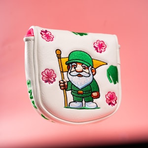 Puede incluir: Una funda blanca para putter de golf con un gnomo bordado que viste traje y gorro verdes, sosteniendo una bandera amarilla. El dise&ntilde;o incluye detalles de flores rosas y texto verde en el lateral, con un forro interior rosa brillante.