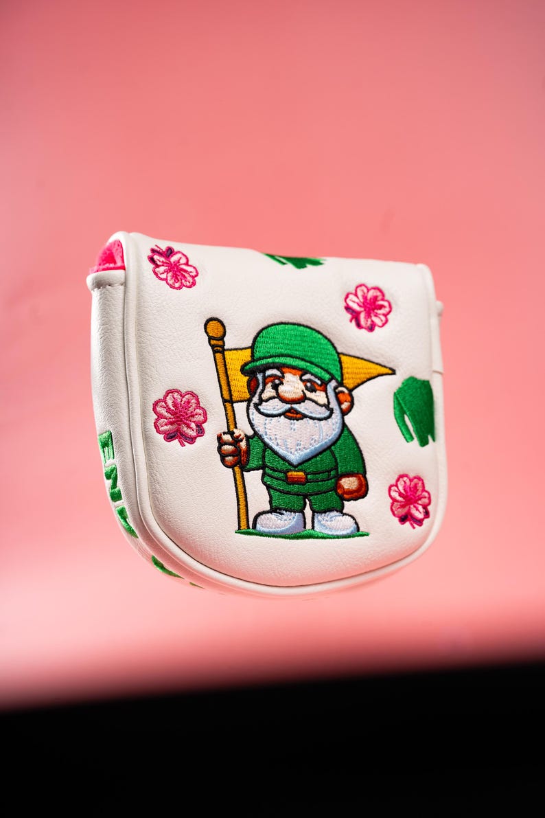 Puede incluir: Una funda blanca para putter de golf con un gnomo bordado que viste traje y gorro verdes, sosteniendo una bandera amarilla. El dise&ntilde;o incluye detalles de flores rosas y texto verde en el lateral, con un forro interior rosa brillante.