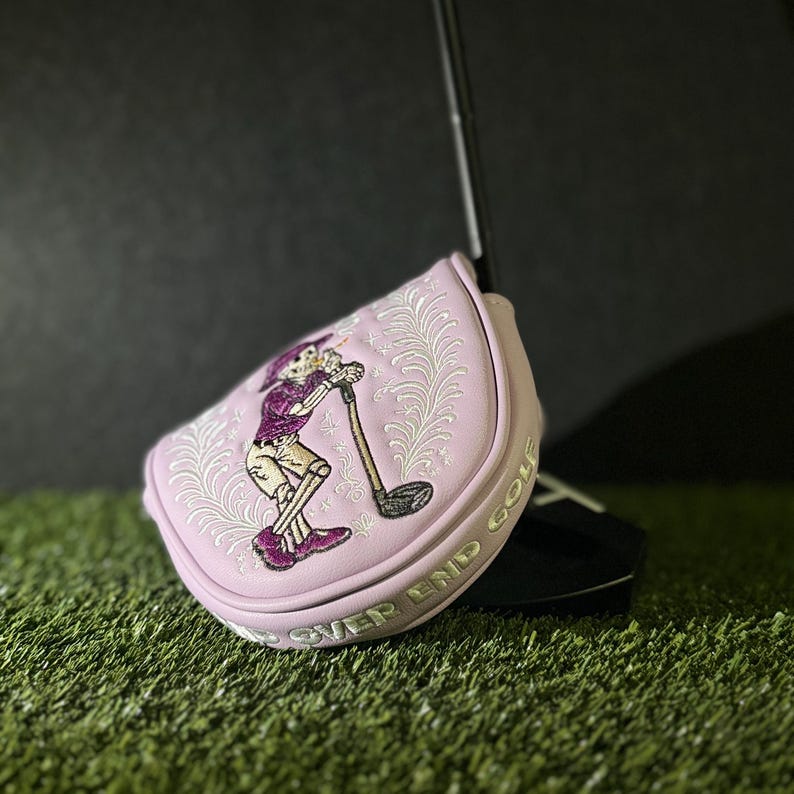 May include: A light pink golf club head cover with an embroidered skeleton design. The skeleton is wearing a purple shirt and shorts, holding a golf club. The cover has the words "OVER END GOLF" in silver lettering.