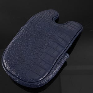 May include: A dark navy blue protective case featuring a textured crocodile skin pattern. The case has a rounded rectangular shape with a distinct U-shaped cutout at the top edge and stitched borders along the entire perimeter of the item.