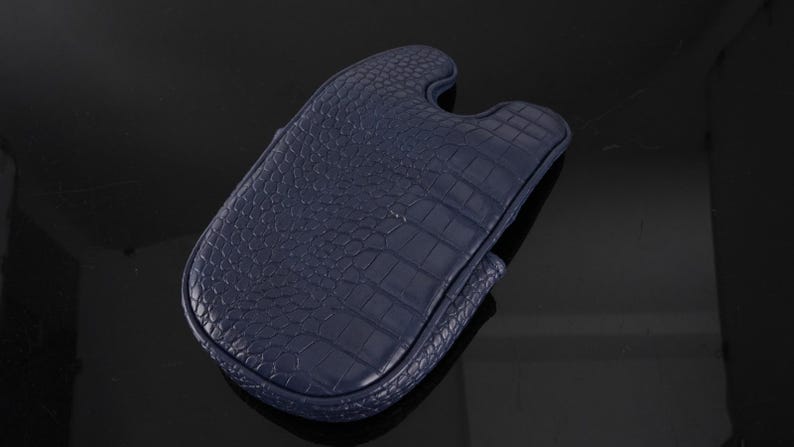 May include: A dark navy blue protective case featuring a textured crocodile skin pattern. The case has a rounded rectangular shape with a distinct U-shaped cutout at the top edge and stitched borders along the entire perimeter of the item.