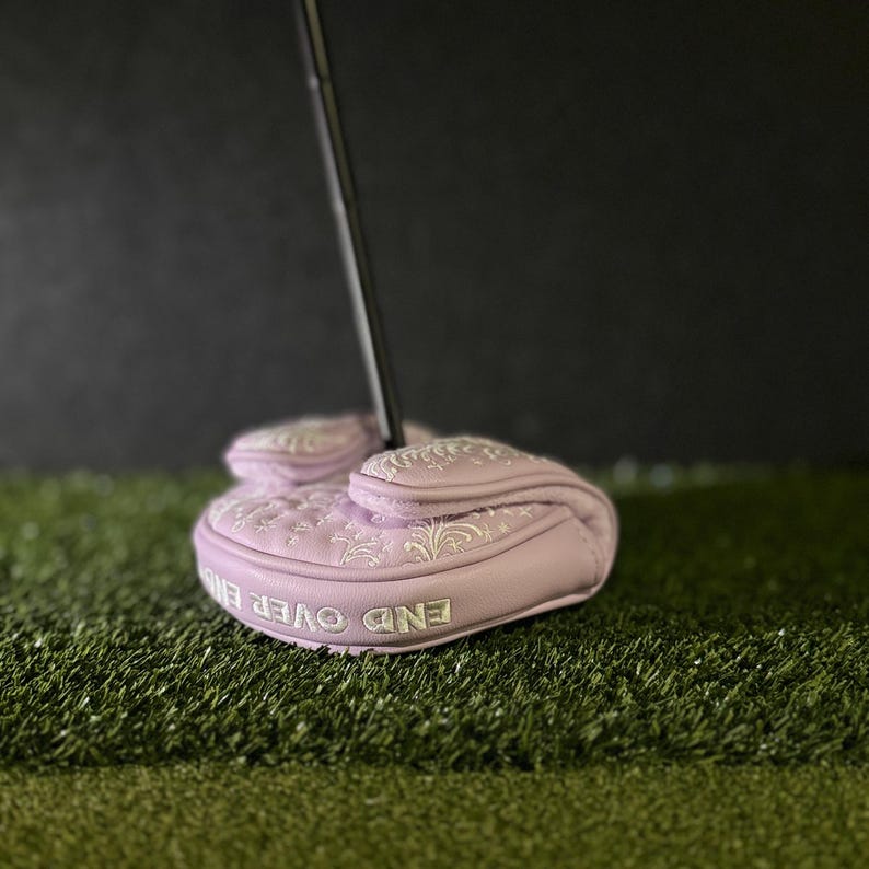 May include: A light purple golf putter head cover with white embroidered floral designs. The cover has the words "END OVER END" in white lettering. The putter is on a green surface.