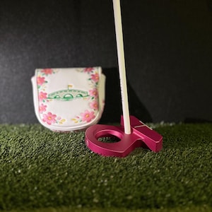May include: A magenta golf putter with a white shaft is set on artificial turf. A white golf club cover with a floral design and a green bridge is to the left. The background is black.