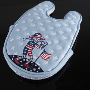 May include: Light blue golf club head cover with white stars and a patriotic skeleton golfer design. The skeleton is wearing a hat and shorts in red, white, and blue, holding a golf club and waving an American flag.