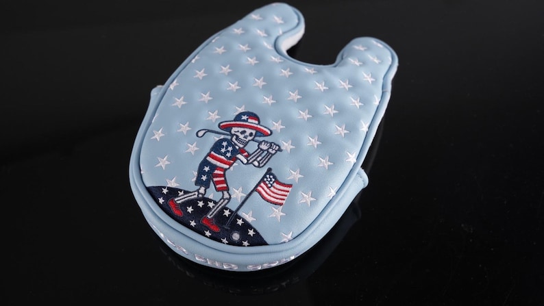 May include: Light blue golf club head cover with white stars and a patriotic skeleton golfer design. The skeleton is wearing a hat and shorts in red, white, and blue, holding a golf club and waving an American flag.