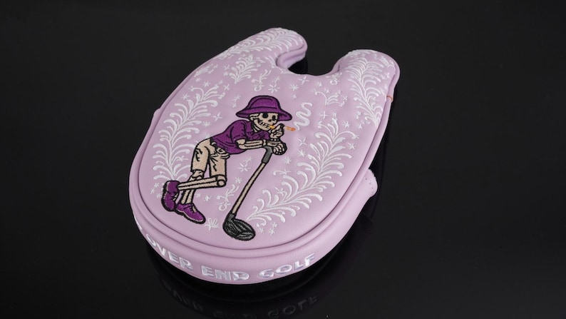 May include: A light purple golf club head cover featuring a cartoon skeleton in a purple hat and shirt, leaning on a golf club. The cover has white floral designs and the words "NEVER END GOLF".