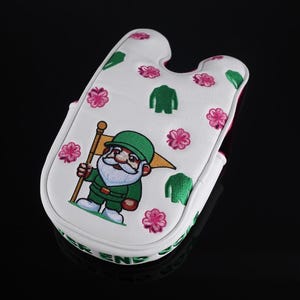 Puede incluir: Una funda para putter de golf blanca con un gnomo bordado que lleva un sombrero y un traje verdes, sosteniendo una bandera amarilla. El dise&ntilde;o incluye patrones de flores rosas e iconos de chaquetas verdes, con texto verde en el borde.