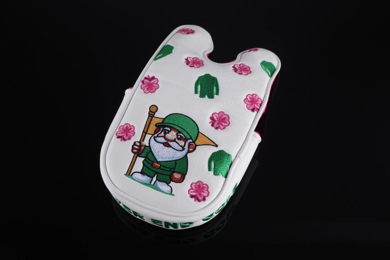 Puede incluir: Una funda para putter de golf blanca con un gnomo bordado que lleva un sombrero y un traje verdes, sosteniendo una bandera amarilla. El dise&ntilde;o incluye patrones de flores rosas e iconos de chaquetas verdes, con texto verde en el borde.