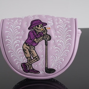 May include: A pale purple golf club head cover featuring an embroidered skeleton in a purple shirt and hat, leaning on a golf club. The cover has a floral design and a curved shape.