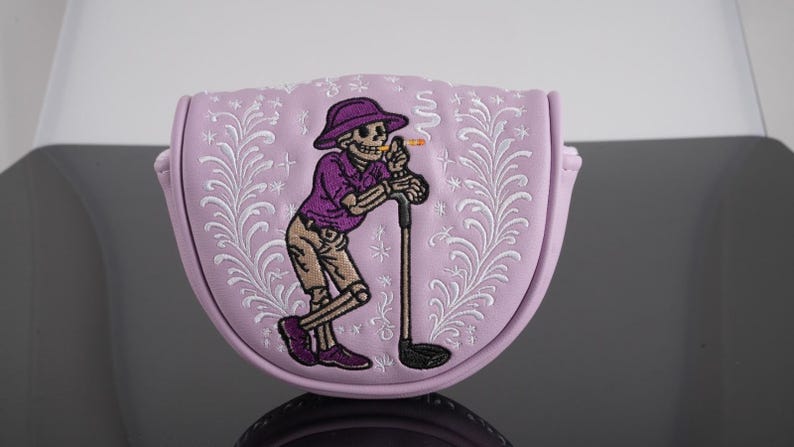 May include: A pale purple golf club head cover featuring an embroidered skeleton in a purple shirt and hat, leaning on a golf club. The cover has a floral design and a curved shape.
