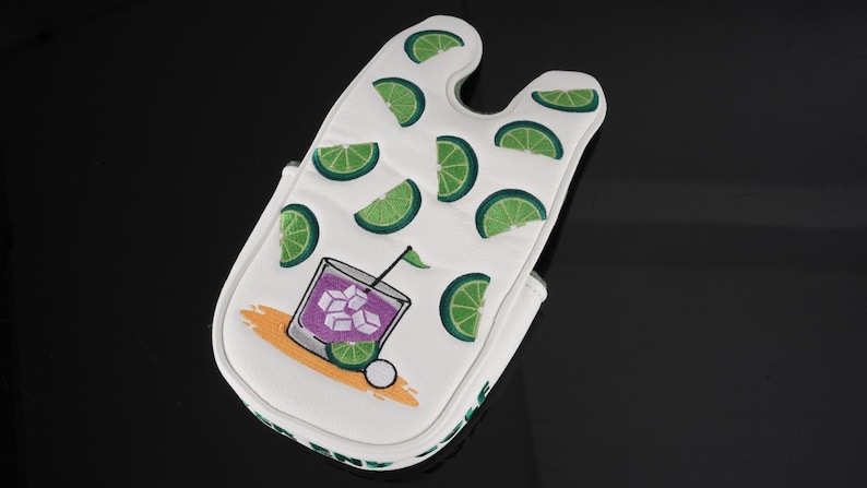 Pu&ograve; includere: Un copri bastone da golf bianco con un motivo ripetuto di fette di lime verde e una grafica di un cocktail viola in un bicchiere con cubetti di ghiaccio, una cannuccia nera e una fetta di lime, su un design a spruzzo arancione chiaro.