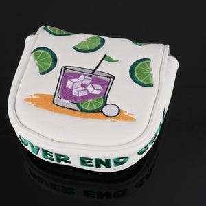 Pu&ograve; includere: Un copri-putter a martello bianco con un ricamo raffigurante un cocktail viola con cubetti di ghiaccio e spicchi di lime. Il design include fette di lime sparse e la scritta OVER END GOLF ricamata in verde lungo il bordo laterale.