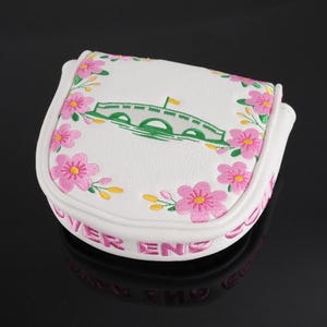 May include: White golf putter cover with pink floral embroidery and a green bridge design. The cover has the words "OVER END" embroidered in pink. A golf accessory.