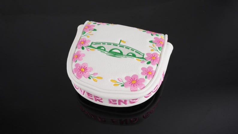 May include: A white mallet-style golf putter headcover featuring an embroidered green bridge graphic, pink cherry blossom flowers, and the text "OVER END" in pink lettering along the side, constructed from a textured synthetic material.