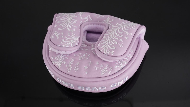 May include: A lilac golf putter cover featuring white embroidered floral patterns. The cover is rounded with a central opening and a curved top. The words "END OVER END" are embroidered along the bottom edge.