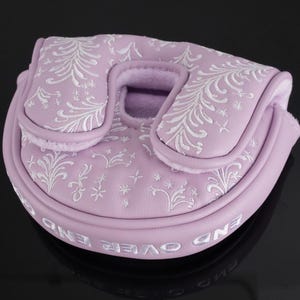 May include: A lavender golf putter cover with white embroidered floral patterns. The cover has a rounded shape with a central opening and a curved top. The words "END OVER END" are embroidered in white around the bottom edge.