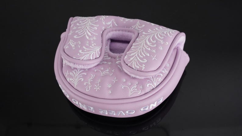 May include: A lavender golf putter cover with white embroidered floral patterns. The cover has a rounded shape with a central opening and a curved top. The words "END OVER END" are embroidered in white around the bottom edge.