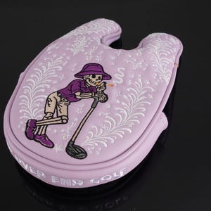 May include: A light purple golf club head cover with a cartoon skeleton golfer design. The skeleton is wearing a purple hat and shirt, leaning on a golf club. The cover has white floral accents and the words "NEVER END GOLF".