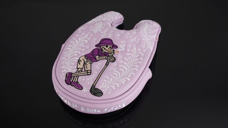 May include: A light purple golf club head cover with a cartoon skeleton golfer design. The skeleton is wearing a purple hat and shirt, leaning on a golf club. The cover has white floral accents and the words "NEVER END GOLF".