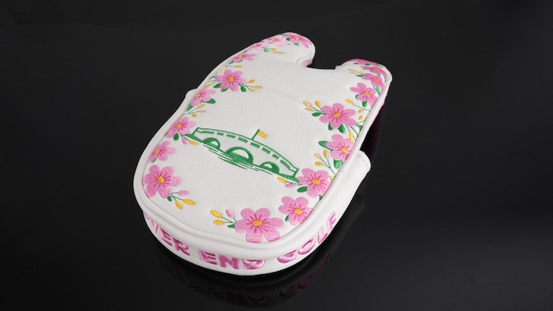 May include: A white golf putter headcover featuring a green bridge illustration and pink floral embroidery. The text "RIVER END GOLF" is embroidered in pink along the side edge. The cover has a U-shaped cutout at the top and a textured surface.