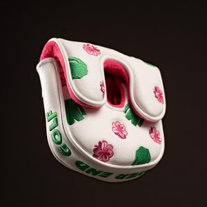 Puede incluir: Una funda blanca para putter de golf con forro interior rosa. El exterior est&aacute; decorado con formas de tr&eacute;boles verdes y patrones de flores rosas bordados. El lateral de la funda muestra el texto bordado en verde "EVER END GOLF".