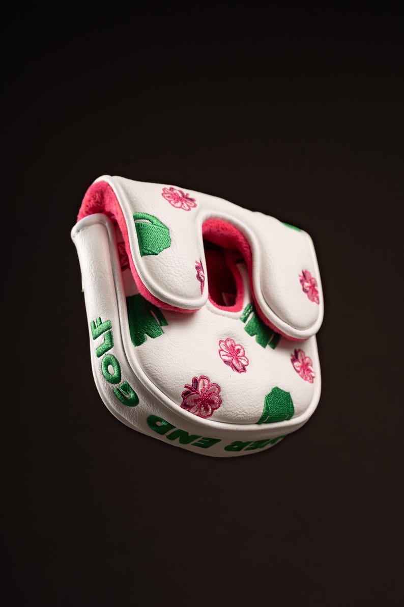 Puede incluir: Una funda blanca para putter de golf con forro interior rosa. El exterior est&aacute; decorado con formas de tr&eacute;boles verdes y patrones de flores rosas bordados. El lateral de la funda muestra el texto bordado en verde "EVER END GOLF".
