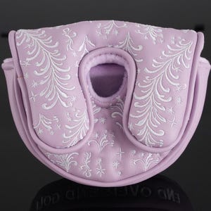 May include: A lavender golf putter cover with white embroidered floral and star patterns. The cover has a rounded shape with a central opening and a magnetic closure. The cover is made of a soft, padded material.