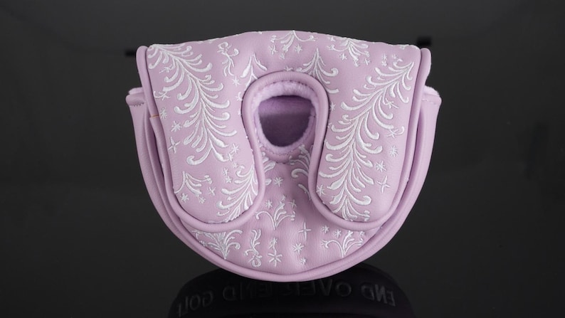 May include: A lavender golf putter cover with white embroidered floral and star patterns. The cover has a rounded shape with a central opening and a magnetic closure. The cover is made of a soft, padded material.