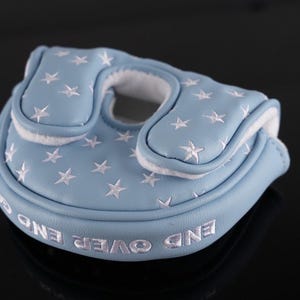 May include: A light blue golf putter cover with white star embroidery. The cover has a white interior and the words "END OVER END" embroidered in white. The cover is made of a soft material and is designed to protect the putter head.