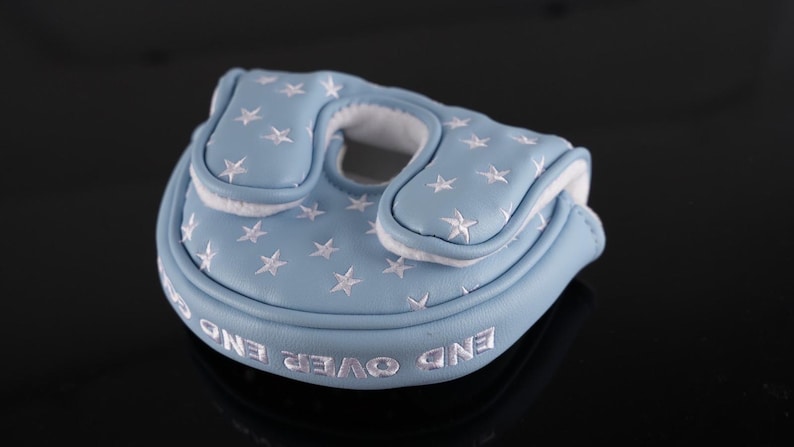 May include: A light blue golf putter cover with white star embroidery. The cover has a white interior and the words "END OVER END" embroidered in white. The cover is made of a soft material and is designed to protect the putter head.