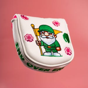 Puede incluir: Una funda blanca para putter de golf con un gnomo bordado vestido de verde que sostiene una bandera amarilla. El dise&ntilde;o incluye detalles de flores rosas y el texto bordado en verde END OVER END a lo largo del borde lateral de la funda.